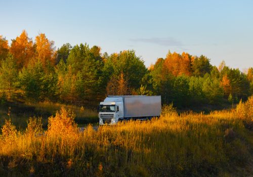 A truck of an unknown brand is driving on a road in an autumnal landscape bathed in the light of the setting sun. The autumn colors, the romance of a long-haul trucker, and the delivery of goods on land. High quality photo